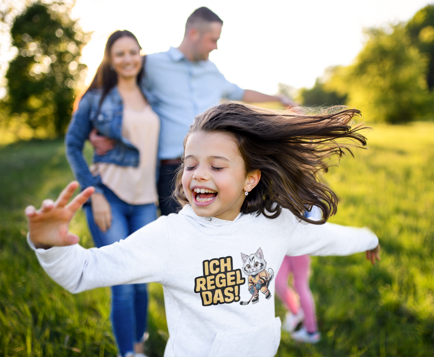 lachendes über eine Wiese rennendes Kind im weißem Pullover mit Aufdruck einer Katze im Eishockeyoutfit und dem Satz ich Regel das! Eltern im Hintergrund.